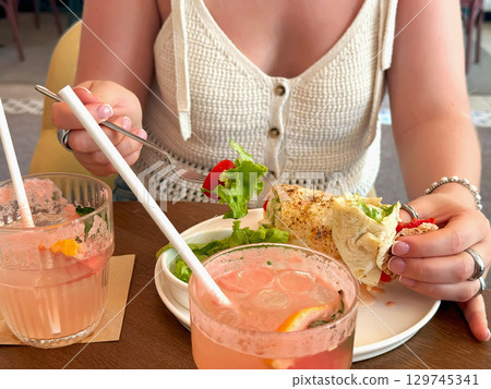 Young woman eats tortilla with chicken and vegetables. Young woman enjoying fresh wrap with salad and cold drink at stylish cafe table. Concept of healthy lunch and summer lifestyle 129745341