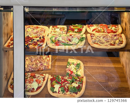 Display of rectangular pizzas with various toppings inside glass showcase at deli. Concept of pizza display, city fast food, quick lunch option 129745366