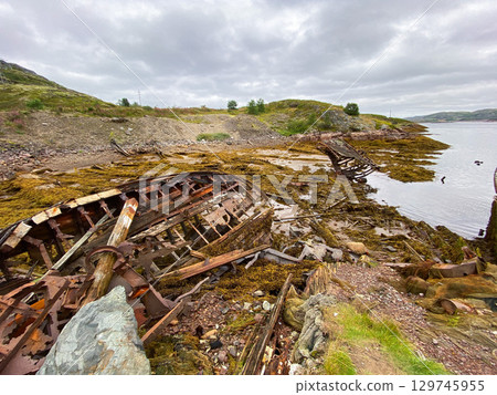 Decaying wooden shipwreck remains on rocky shore under cloudy mountain sky. 129745955