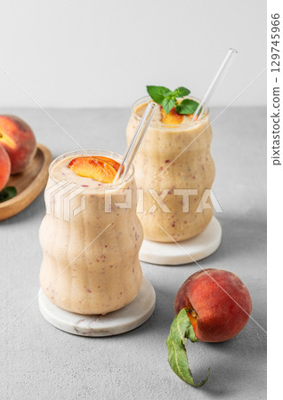 Two glasses of smoothie with peach and banana in a glass on a light background with tropical fruits. 129745966