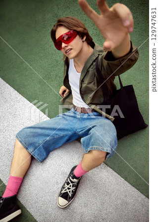 Young man in streetwear and red glasses sitting on ground reaching to camera 129747631