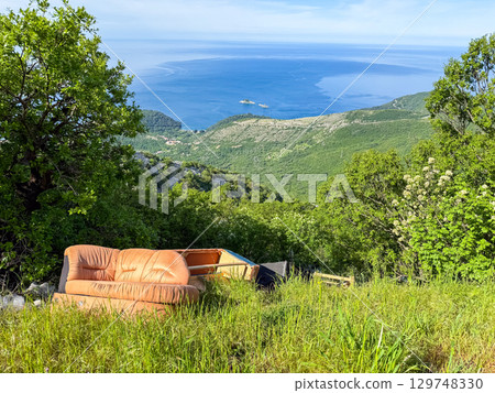 Old sofa abandoned on green hillside with panoramic sea view. Contrast of natural serenity and human neglect in a wild coastal landscape. Copy space 129748330