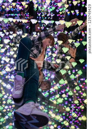 Child lying on reflective surface, engaging with smartphone near vibrant LED light installation, displaying modern digital interaction Child lying on reflective surface, engaging with smartphone near vibrant LED light installation, displaying modern digital interaction 129749429