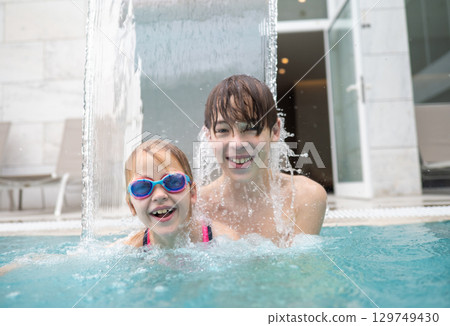 Happy brother and sister enjoying playful moments in a swimming pool, splashing under a refreshing waterfall on a sunny summer day Happy brother and sister enjoying playful moments in a swimming pool, splashing under a refreshing waterfall on a sunny summer day 129749430