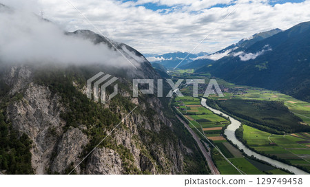 Aerial view of a valley in the Austrian Alps with low clouds covering the mountainside, featuring the Inn river, fields, and a small town Aerial view of a valley in the Austrian Alps with low clouds covering the mountainside, featuring the Inn river, fields, and a small town 129749458