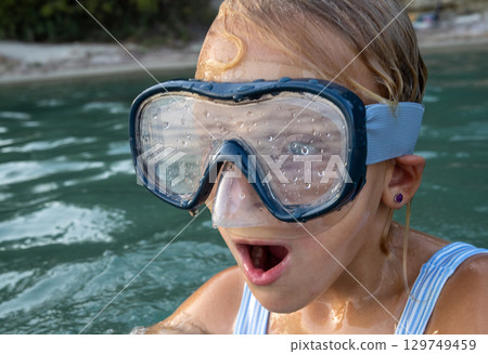 Portrait of a surprised girl wearing a diving mask, swimming joyfully in the sea during a fun filled summer vacation by the beach 129749459