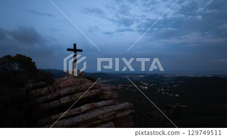 Large cross stands tall on rocky terrain. Casting a striking silhouette against the darkening sky. With a glowing city in the distance and drifting clouds. Evoking hope and spiritual contemplation 129749511