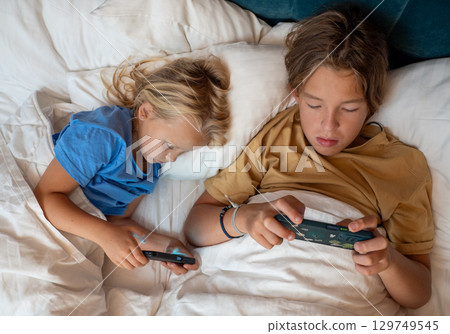 Two young siblings lying under a cozy blanket, playing video games on their smartphones while enjoying a relaxed morning in bed Two young siblings lying under a cozy blanket, playing video games on their smartphones while enjoying a relaxed morning in bed 129749545