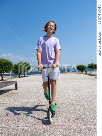 Carefree young boy riding a scooter through a vibrant park by the sea, soaking up the sun and enjoying a joyful summer day 129749546