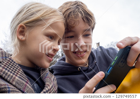 Siblings enjoying a fun outdoor gaming session on their smartphones, sharing smiles and laughter under a cloudy sky 129749557