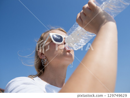 Woman with sunglasses drinking bottled water, basking in bright sunlight against cloudless blue sky during summer outdoor scene Woman with sunglasses drinking bottled water, basking in bright sunlight against cloudless blue sky during summer outdoor scene 129749629