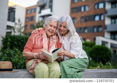 Elderly mother sitting on bench with adult daughter, reading book. 129750701