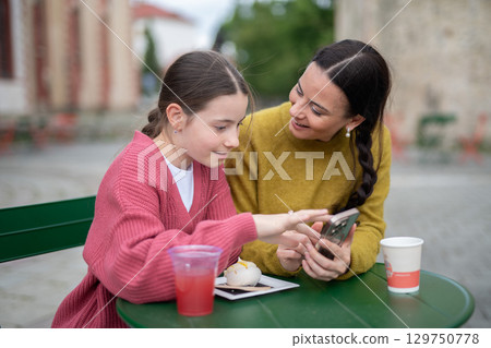 Mother and daughter talking over drink and dessert, looking at smartphone screen. 129750778