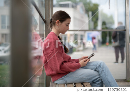 Young girl sitting at bus stop, scrolling through social media feed. Young girl sitting at bus stop, scrolling through social media feed. 129750801