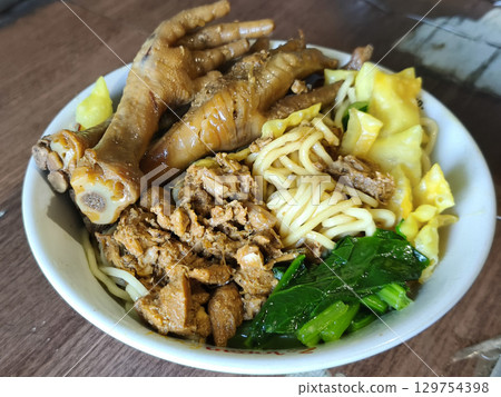 A close-up shot of a bowl of Mie Ayam or Indonesian style chicken noodles with chicken feet, minced chicken, and vegetables on a wooden table. A close-up shot of a bowl of Mie Ayam or Indonesian style chicken noodles with chicken feet, minced chicken, and vegetables on a wooden table. 129754398