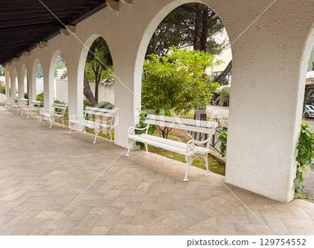 White benches under arched walkway with garden view. Architecture, relaxation, and peaceful public space for leisure. 129754552
