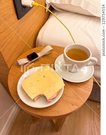 Cup of tea with sandwich and smartwatch on bedside table. Morning routine, casual breakfast, and modern lifestyle at home. 129754554