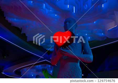 Futuristic art installation with glowing red heart held by humanoid statue under dramatic blue neon lights and abstract ceiling design. Modern statue with the concept of love and kindness 129754676