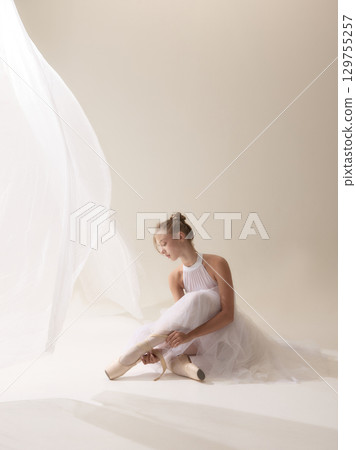 Time between movements. Young ballerina adjusting ribbons of pointe shoes in soft light 129755257