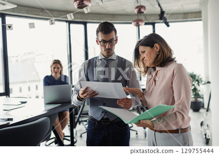 Two collegues talking about project document, standing in modern office. 129755444