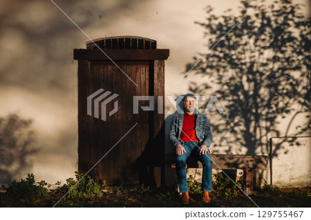 Older man enjoying warm autumn sun in face, sitting on bench. 129755467