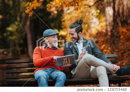 Senior father and his son sitting on bench by lake in nature, looking at tablet. Senior father and his son sitting on bench by lake in nature, looking at tablet. 129755480