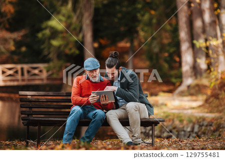 Senior father and his son sitting on bench by lake in nature, looking at tablet. 129755481