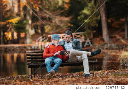 Senior father and his son sitting on bench by lake in nature, looking at tablet. Senior father and his son sitting on bench by lake in nature, looking at tablet. 129755482