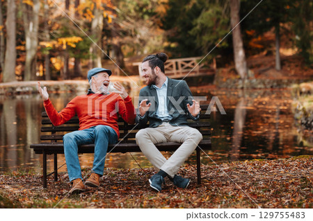 Grandad and grandson sitting on bench by lake and talking. 129755483