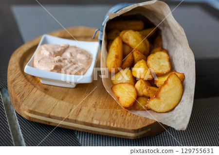 Rustic Fried potato wedges in a paper envelope with sauce on a wooden plate on the outdoor terrace in a cafe 129755631