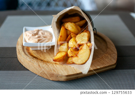 Rustic Fried potato wedges in a paper envelope with sauce on a wooden plate on the outdoor terrace in a cafe 129755633