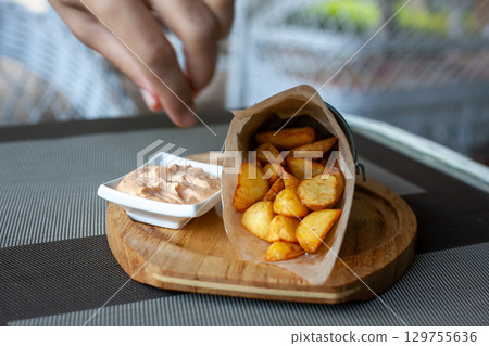 Rustic Fried potato wedges in a paper envelope with sauce on a wooden plate on the outdoor terrace in a cafe. Mans hand takes slice and drops it into sauce 129755636