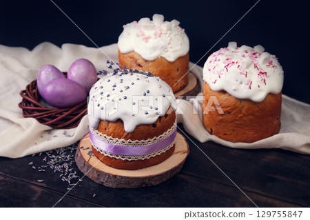Easter cakes on a dark wooden background, decorated with lavender and marshmallows, and painted Easter eggs in a wicker nest. 129755847