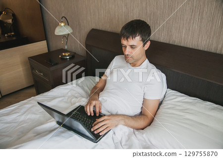 A middle-aged man is sitting in bed in his bedroom and working on a laptop 129755870