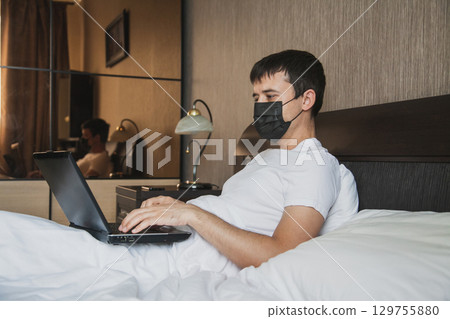 A man is sitting in bed in his bedroom and working at a laptop with a medical mask on his face A man is sitting in bed in his bedroom and working at a laptop with a medical mask on his face 129755880