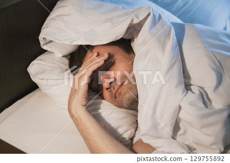 Man experiencing discomfort in bed while covered in blankets in a dimly lit bedroom during morning hours 129755892