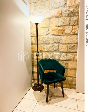 A green velvet chair with a tall lamp stands in the stone wall corner indoors. Interior design, hospitality and cozy atmosphere expressed through modern lounge furniture. A green velvet chair with a tall lamp stands in the stone wall corner indoors. Interior design, hospitality and cozy atmosphere expressed through modern lounge furniture. 129757240
