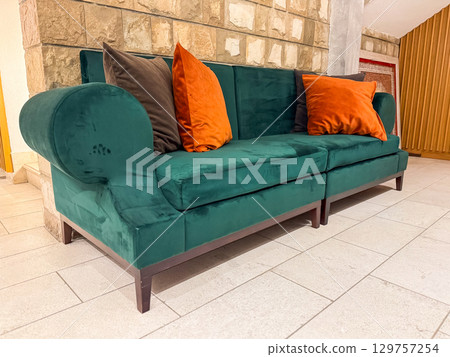 A green velvet sofa with bright orange pillows stands against a stone wall interior. Comfort, style and modern hospitality expressed through colorful lounge furniture. A green velvet sofa with bright orange pillows stands against a stone wall interior. Comfort, style and modern hospitality expressed through colorful lounge furniture. 129757254