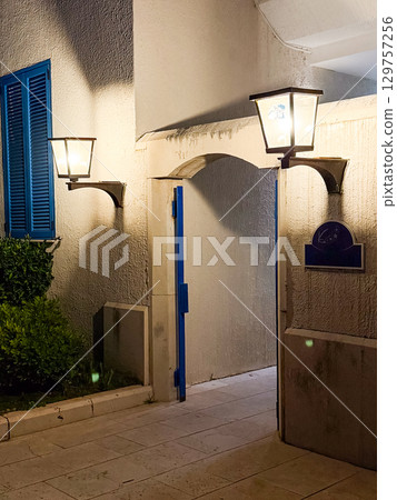 An illuminated entrance with blue shutters and open doorway shines under two glowing lanterns. Tradition, hospitality and evening atmosphere expressed through rustic architecture. An illuminated entrance with blue shutters and open doorway shines under two glowing lanterns. Tradition, hospitality and evening atmosphere expressed through rustic architecture. 129757256
