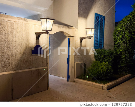 An illuminated entrance with blue shutters and open doorway shines under two glowing lanterns. Tradition, hospitality and evening atmosphere expressed through rustic architecture. An illuminated entrance with blue shutters and open doorway shines under two glowing lanterns. Tradition, hospitality and evening atmosphere expressed through rustic architecture. 129757258