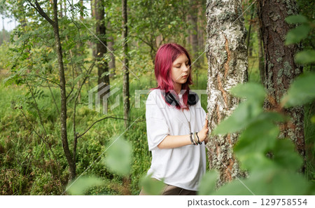 Young girl with pink hair exploring forest and touching tree trunk 129758554