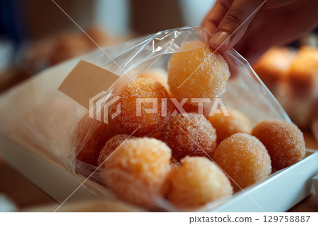 Sugary round donuts in a clear plastic bag being picked up by hand, displayed in a white box in warm natural lighting 129758887