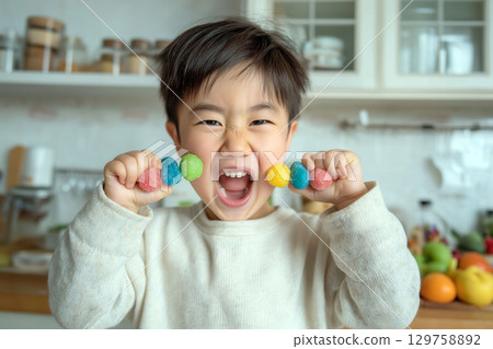 Excited young boy holding two colorful candy skewers and smiling widely in a bright home kitchen setting 129758892