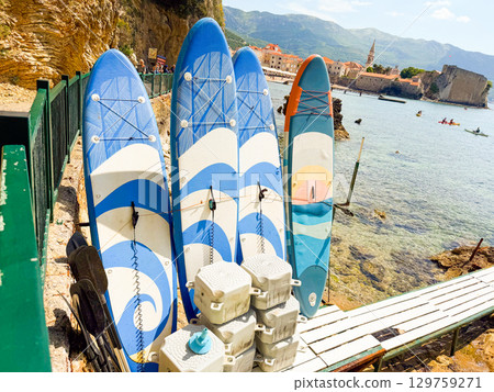 Surfboards with paddles placed by the sea with town and mountains in background. Adventure, recreation, summer lifestyle and coastal tourism in seaside travel scene. Surfboards with paddles placed by the sea with town and mountains in background. Adventure, recreation, summer lifestyle and coastal tourism in seaside travel scene. 129759271