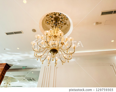 A golden chandelier with crystal ornaments hanging from a decorated ceiling. Elegance, luxury, atmosphere and architectural style in hotel interior and dining hall design. A golden chandelier with crystal ornaments hanging from a decorated ceiling. Elegance, luxury, atmosphere and architectural style in hotel interior and dining hall design. 129759303