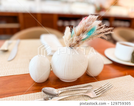 A ceramic vase with dried plants and tableware on a decorated dining table. Interior, detail, elegance and hospitality in restaurant atmosphere and dining experience. A ceramic vase with dried plants and tableware on a decorated dining table. Interior, detail, elegance and hospitality in restaurant atmosphere and dining experience. 129759305