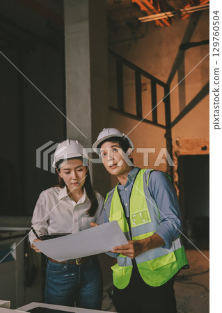 Construction manager and engineer dressed in orange work vests and hard helmets explore construction documentation on the building site near the steel frames 129760504