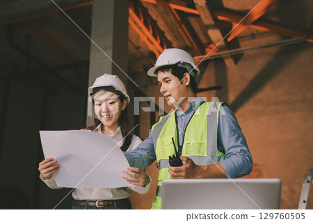 Construction manager and engineer dressed in orange work vests and hard helmets explore construction documentation on the building site near the steel frames 129760505