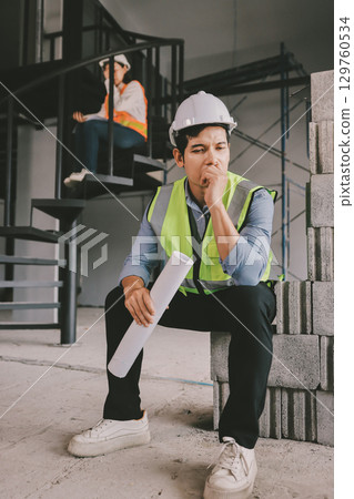 Construction manager and engineer dressed in orange work vests and hard helmets explore construction documentation on the building site near the steel frames 129760534