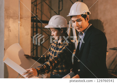 Construction manager and engineer dressed in orange work vests and hard helmets explore construction documentation on the building site near the steel frames Construction manager and engineer dressed in orange work vests and hard helmets explore construction documentation on the building site near the steel frames 129760541